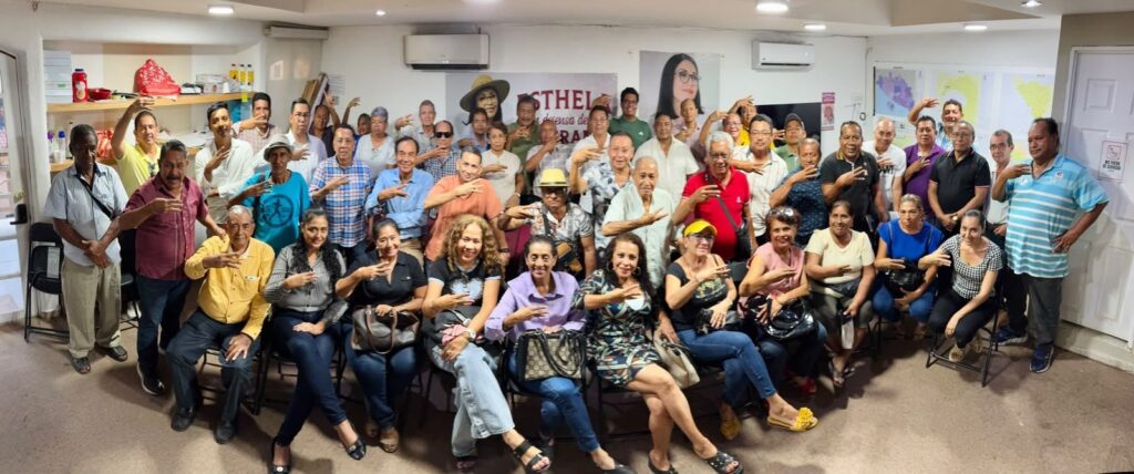 Foto grupal de una reunión numerosa con asistentes saludando con la mano en alto